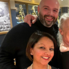 The Retail Partnerships team posing together in a warmly lit setting with festive lights in the background. A smiling woman with short brown hair, pearl earrings, and a pearl necklace is in the foreground. Behind her, a bald man with a beard, wearing a black sweater. The setting is in a  restaurant with framed artwork on the walls and decorated Christmas trees reflecting in the windows.