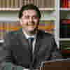 Tommy Salisbury smiles at the camera at a table in an office, he is in front of a shelf of books and in front of him a laptop. 