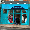 A vibrant blue storefront with signage reading 'Fight for Sight' and 'Save Sight. Change Lives.' The shop, located at 124A, has large glass windows displaying clothing, accessories, and festive decorations. A person wearing a blue and black jacket, jeans, and bright orange sneakers walks past the store with a black dog.