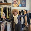 Volunteer Ada in our West Norwood charity shop - Ada has her arms spread wide and a big smile on her face, she is standing in front of rails of colourful clothing