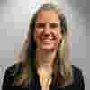 Kim has shoulder-length grey hair and smiles warmly in a medium-shot portrait. The image presents a straightforward portrait of a woman, centered within the frame. The woman's expression is one of warmth and friendliness. The lighting is even and highlights her features without harsh shadows.