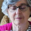 Susan, a woman with white hair and wearing glasses is concentrating and looking down at something in her hands. She's wearing a pink cardigan and sitting in an armchair