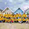 A large group of young adults all wearing yellow hoodies are posing on a sandy beach in front of a row of colourful beach huts. Some of the people have their arms around each other's shoulders, some are kneeling on the sand. They are all looking very happy